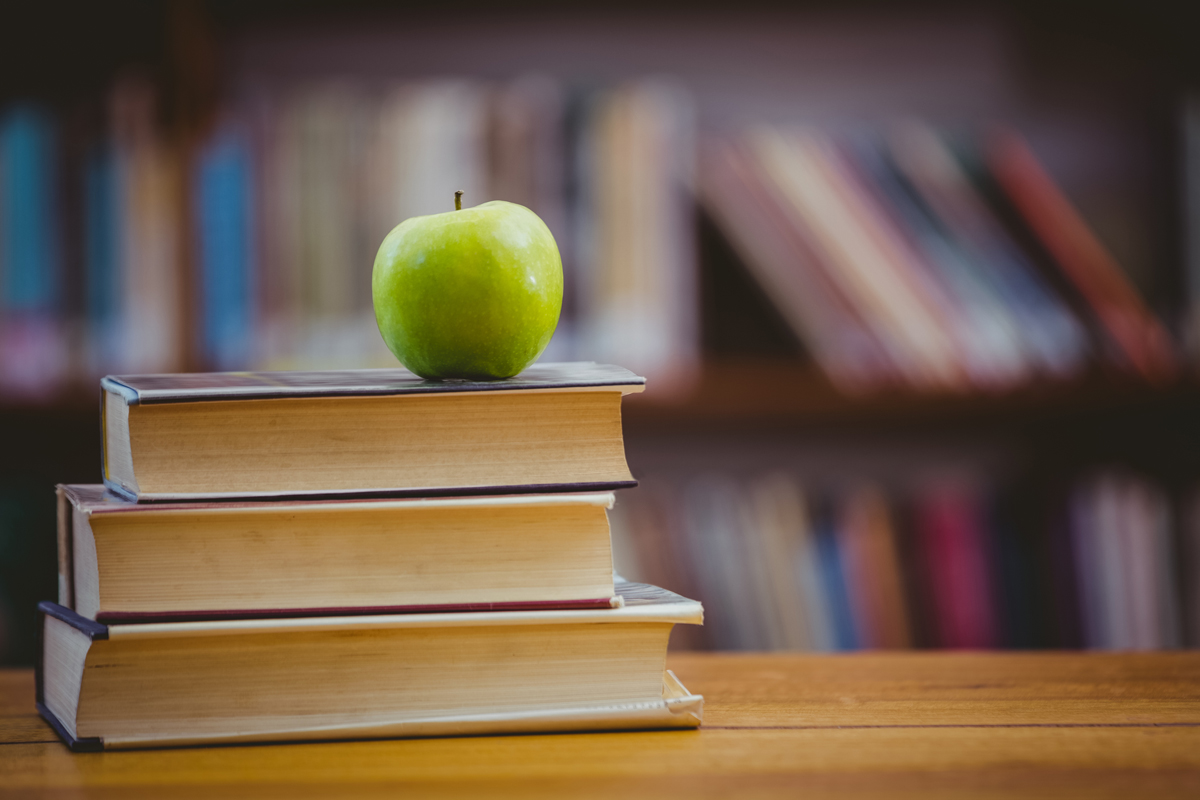 An apple sits on a pile of books with a library shelf in the background