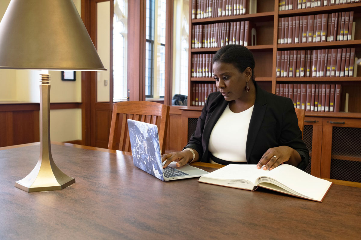 A graduate student studies in the TC library using a book and her laptop.